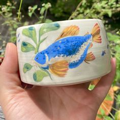 a hand is holding a blue and yellow fish on a white ceramic container in front of some plants