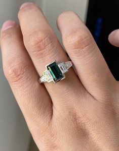 a woman's hand holding a ring with an emerald and diamond set in it