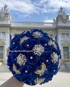 a bridal bouquet with blue flowers and gold brooches in front of a building