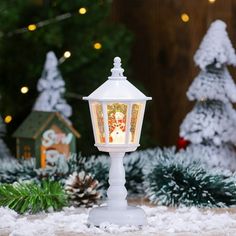 a white lantern with a christmas scene on it in front of snow covered evergreen trees