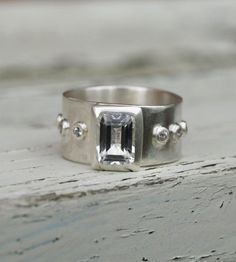 A 10mm wide silver band is set with an emerald cut white topaz. Around the band are six small moissanites also set in silver.This ring is in stock and ready to ship it will fit a size 7 ½-8 r a size P.The ring could also be made in other metals just ask for a quote.For other white topaz rings go to:www.etsy.com/shop/karenjohnsondesignorfacebook.com/karenjohnsondesigninstagram.com/karenjohnsondesign Modern Silver Topaz Ring With Emerald Cut, White Topaz Jewelry With Emerald Cut, White Topaz Emerald-cut Jewelry, White Topaz Emerald Cut Jewelry, White Emerald Cut Topaz Jewelry, Wide Silver Band, Topaz Rings, Topas Ring, Sterling Silver Jewelry Rings