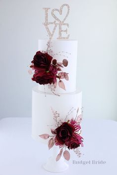 two tiered wedding cake with red flowers and love spelled on the top, sitting on a white table