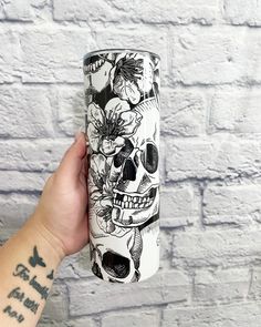 a hand holding up a white and black coffee cup with skulls on it, flowers in the background