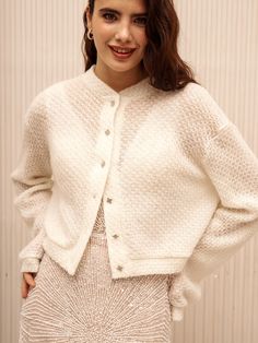 a woman standing in front of a wall wearing a white cardigan sweater and skirt