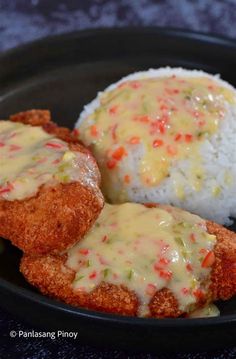 two pieces of fried chicken covered in gravy and rice on a black plate