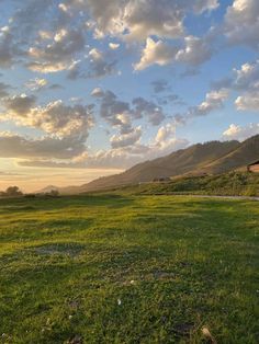 the sun is setting over a grassy field with mountains in the background and clouds overhead