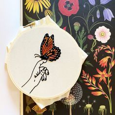 a hand embroidered butterfly sitting on top of a white piece of fabric next to a floral wallpaper