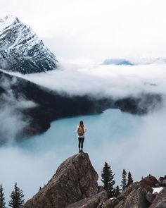 a woman standing on top of a mountain with her hands in her pockets looking at the clouds