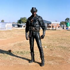 a man in black leather is standing on the dirt field with his hands behind his back