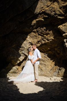 a bride and groom standing in front of a rock formation on the beach at sunset