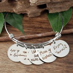 three silver bracelets with handwritten words on them sitting on a wooden table next to leaves