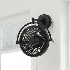 a black wall mounted fan hanging from the side of a white wall next to a window