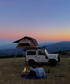 two people sitting in the grass near a camper with a tent on its roof