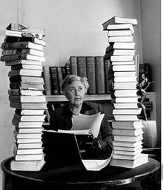 an old woman sitting at a table with stacks of books on top of her head