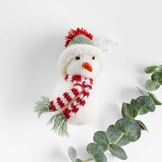 a knitted snowman ornament hanging from a green leafy branch on a white background