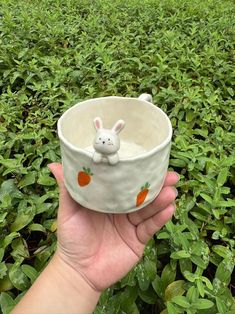 a hand holding a bowl with a small bunny in it on top of some bushes