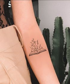 a person with a tattoo on their arm holding an open book in front of a cactus