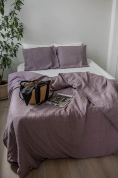 a bed with purple sheets and a black purse on top of it in front of a white wall