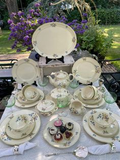 a table with plates and cups on it in the middle of a garden area, surrounded by purple flowers