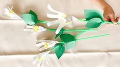 a person is holding some paper flowers on a white sheet with green stems and leaves