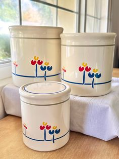 two white containers sitting on top of a table next to a window with flowers painted on them