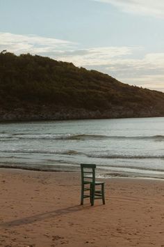 a green chair sitting on top of a sandy beach next to the ocean with a hill in the background