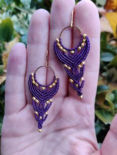 a pair of purple and gold beaded earrings on a person's hand in front of green leaves