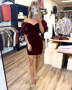 a woman taking a selfie in front of a clothing store mirror wearing a burgundy velvet dress