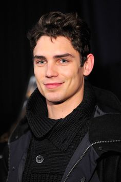 a young man in a black jacket and scarf smiles at the camera while looking into the camera