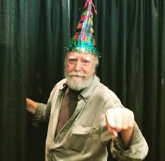 an older man wearing a party hat pointing at the camera while standing in front of a black curtain