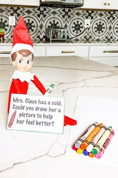 an elf holding a sign next to some crayons on the kitchen counter top