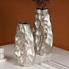 two silver vases sitting on top of a white table next to a planter