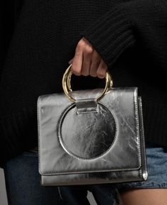 The Hunter is a sleek new addition to the Alexis Bittar handbag line, mixing 70's minimalism with a modern approach. With insets adorned with oversized 14k gold plated rings, inspired by our signature bangles. The rings can be used as top handles or simply ornamental when the bag is converted to a crossbody using the detachable leather strap. Modern Silver Shoulder Bag For Formal Events, Modern Box Shoulder Bag With Metal Hardware, Trendy Metallic Silver Evening Bags, Modern Silver Evening Shoulder Bag, Trendy Metallic Shoulder Bag For Formal Occasions, Modern Silver Bags With Detachable Strap, Modern Silver Bag With Detachable Strap, Modern Silver Shoulder Bag With Top Handle, Modern Silver Shoulder Bag With Detachable Handle