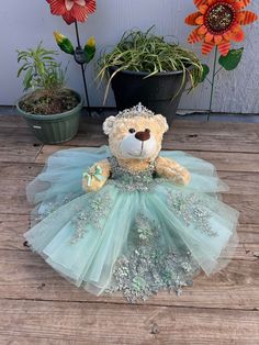 a teddy bear dressed in a dress sitting on a wooden floor next to potted flowers