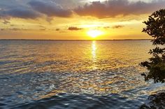 the sun is setting over the water with clouds in the sky and trees on the shore