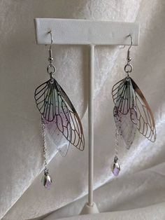 These gorgeous and delicate fairy earrings feature metal and resin wings with a hanging drop crystal. Perfect for special occasions or to add a bit of magic to your every day! The primary color refers to the metal The secondary color refers to the resin wing Silver Fairycore Earrings For Party, Nickel Free Fairycore Dangle Earrings, Nickel-free Fairycore Dangle Earrings, Fairycore Nickel-free Dangle Earrings, Silver Fairy Earrings For Party, Iridescent Fairy Jewelry For Parties, Iridescent Fairy-style Jewelry For Parties, Silver Fairy Style Party Earrings, Iridescent Fairy Style Jewelry For Party