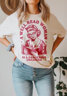 Celebrate the power of knowledge and embrace the spirit of rebellion with this shirt design that boasts a vintage 1950s illustration of a woman immersed in a book. As she captivates the era's charm, the surrounding text boldly declares, "A well-read woman is a dangerous creature." Ideal for book lovers, feminists, and banned book activists, this design encapsulates the strength and influence that come with intellectual prowess.  The juxtaposition of the retro aesthetic with the empowering messag Bookish Crew Neck Shirt With Graphic Print, Relaxed Fit Graphic Print Shirt In Bookish Style, Relaxed Fit Graphic Print Shirt With Bookish Style, Literary Graphic Print Crew Neck T-shirt, Literary Cotton Relaxed Fit T-shirt, Literary Style Cotton T-shirt With Relaxed Fit, Bookish Cotton Shirt With Graphic Print, Literary Cotton T-shirt With Text Print, Bookish Cotton Tops With Character Print