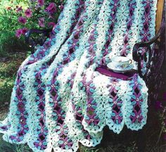 a crocheted blanket sitting on top of a wooden bench next to purple flowers