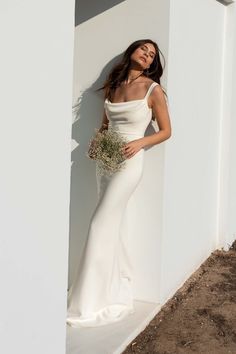 a woman in a long white dress leaning against a wall holding a bouquet and posing for the camera