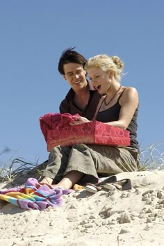 a man and woman sitting on top of a sandy beach next to each other holding blankets