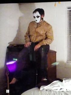 a man sitting on top of a bed wearing a white mask and black leather pants