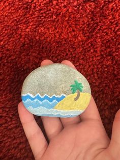 a hand holding a painted rock with a palm tree on it and waves in the sand