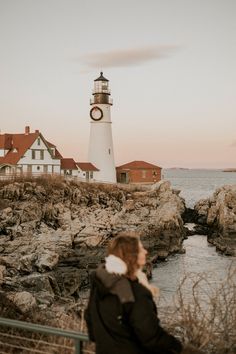 a woman is standing on the rocks looking at the lighthouse