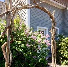 a wooden arch made out of branches in front of a house with bushes and flowers around it