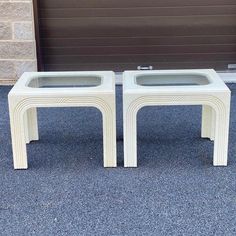 two white side tables with glass top sitting in front of a garage door on asphalt