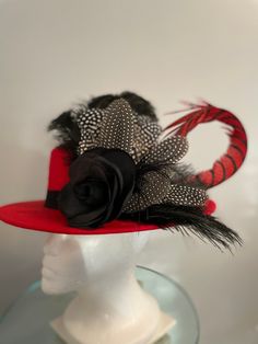 "A red felt \"boater style\" flat crown medium brimmed hat features a large looped red and black Lady Amherst feather, dotted fowl feathers, black ostrich feathers, and a black silk flower that serves as a focal point. The red black and white combo makes this a great Fall hat to wear to the races such as Keeneland, Churchill Downs, carriage driving or any equestrian event. Pair it with a simple black dress and you will turn heads! It is so neutral you can dress it up or down and it will go with just about anything. It's the perfect hat for fall races (Breeders' Cup Nov. 5 at Keeneland) or just wearing on a rainy or cold day. Hatsoffbyhelen.com makes one of kind Hats & Fascinators in Louisville, KY home of the prestigious Kentucky Derby. Each unique piece has its own name resembling a horse Black Top Hat For Kentucky Derby With Flat Crown, Black Top Hat With Flat Crown For Kentucky Derby, Feathered Top Hat For Races, Feathered Top Hat With Short Brim For Races, Feathered Short Brim Top Hat For Races, Feathered Kentucky Derby Hat With Short Brim, Feathered Short Brim Hat For Kentucky Derby, Feathered Costume Hat For Races With Short Brim, Red Western Fedora For Kentucky Derby