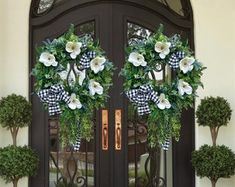 two wreaths on the front door of a house with potted trees and bushes