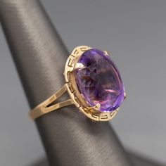 Pretty in purple! This ring features a sparkling oval cut amethyst measuring ~17mm x 14mm, calculating to just over 12ct. The amethyst is a medium purple color with hints of violet and pink. The stone is held with four prongs and surrounded by a frame featuring a greek key border. The ring is not shy, measuring just under 7/8" from front to back. The ring is crafted in 14k yellow gold and weighs 6.57g. It is currently a size 7 and can be resized before shipment for a nominal fee, please ask befo Luxury Purple Amethyst Oval Cabochon Ring, Purple Amethyst Oval Cabochon Ring, Classic Oval Purple Amethyst Ring, Elegant Purple Amethyst Ring Oval Cabochon, Elegant Purple Oval Cabochon Amethyst Ring, Formal Lavender Amethyst Ring With Oval Shape, Formal Lavender Oval Amethyst Ring, Oval Purple Amethyst Ring, Formal Oval Cabochon Amethyst Ring