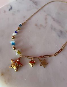 This necklace is a unique and charming piece featuring an eclectic mix of elements. The 21 inch chain is a combination of different sections, with one side adorned with small, colorful stone beads in shades of blue, white, green, and gold, adding a playful touch. The centerpiece of the necklace includes three gold-toned charms: a star with embedded small crystals, a shiny heart, and a delicate flower with cutout petals. The other side of the chain is composed of a more structured, elongated link Bohemian Multicolor Star-shaped Jewelry, Bohemian Charm Necklace With Starfish Charm As Gift, Bohemian Charm Necklace With Starfish For Gift, Bohemian Star-shaped Necklace For Gifts, Bohemian Starfish Charm Necklace For Gift, Bohemian Star Shaped Necklace For Gift, Whimsical Star-shaped Jewelry With Star Charm, Trendy Multicolor Star Charm Necklace, Metal Necklace With Starfish Charm For Gift