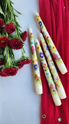 four candles with flowers painted on them sitting next to red cloth and flower bouquets